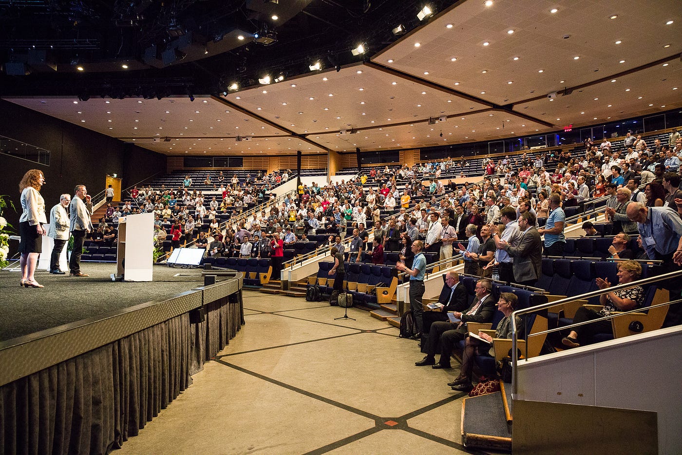 A large academic conference held in a tiered lecture auditorium with presenters on stage.